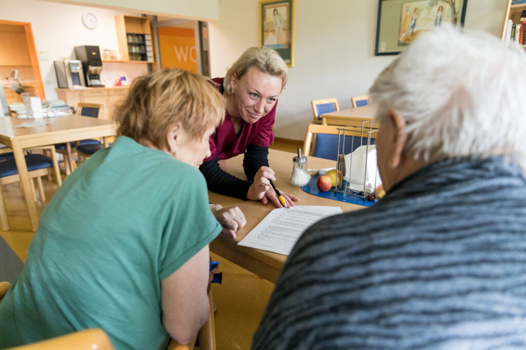 Pflegekraft spricht mit zwei älteren Personen an einem Tisch und erklärt ein Dokument in freundlicher Atmosphäre eines Aufenthaltsraums.