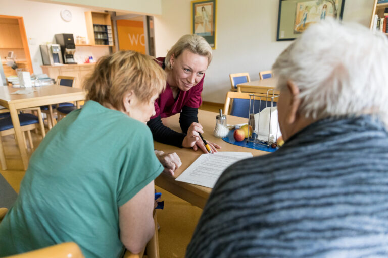 Pflegekraft spricht mit zwei älteren Personen an einem Tisch und erklärt ein Dokument in freundlicher Atmosphäre eines Aufenthaltsraums.