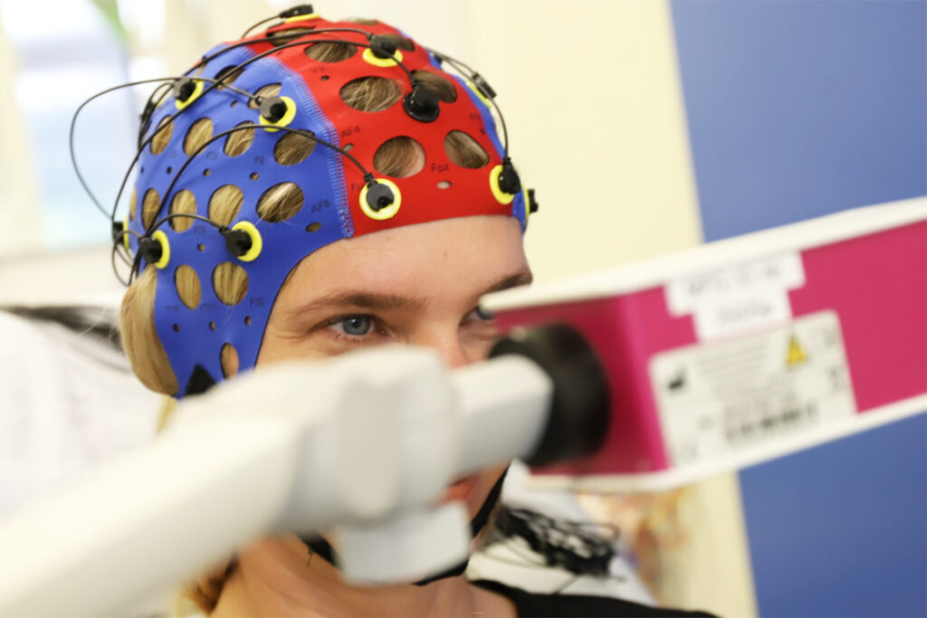 Person trägt eine farbige EEG-Haube mit Elektroden, während sie in ein medizinisches Messgerät blickt.