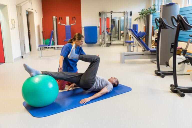 Physiotherapeutin betreut Patienten bei Übung mit Gymnastikball im Therapieraum.