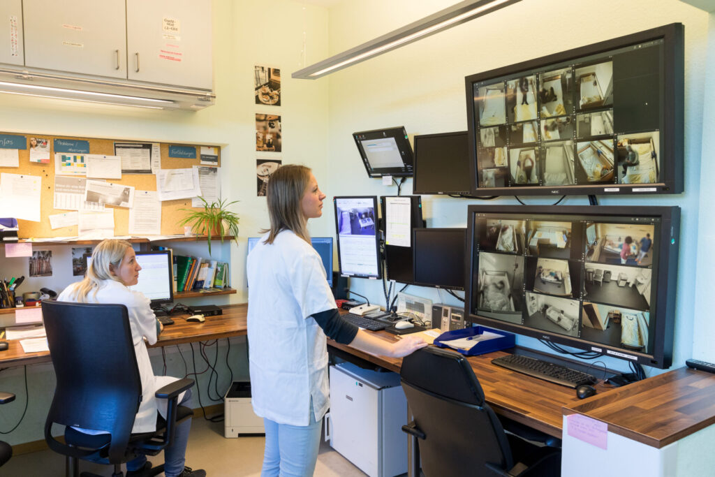 Medizinisches Personal überwacht Patientenräume über mehrere Bildschirme im Kontrollraum.